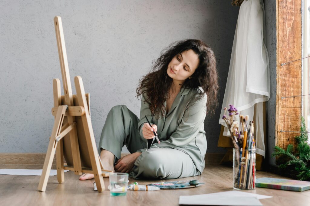 A woman in comfortable attire paints leisurely by a window with art supplies around her.